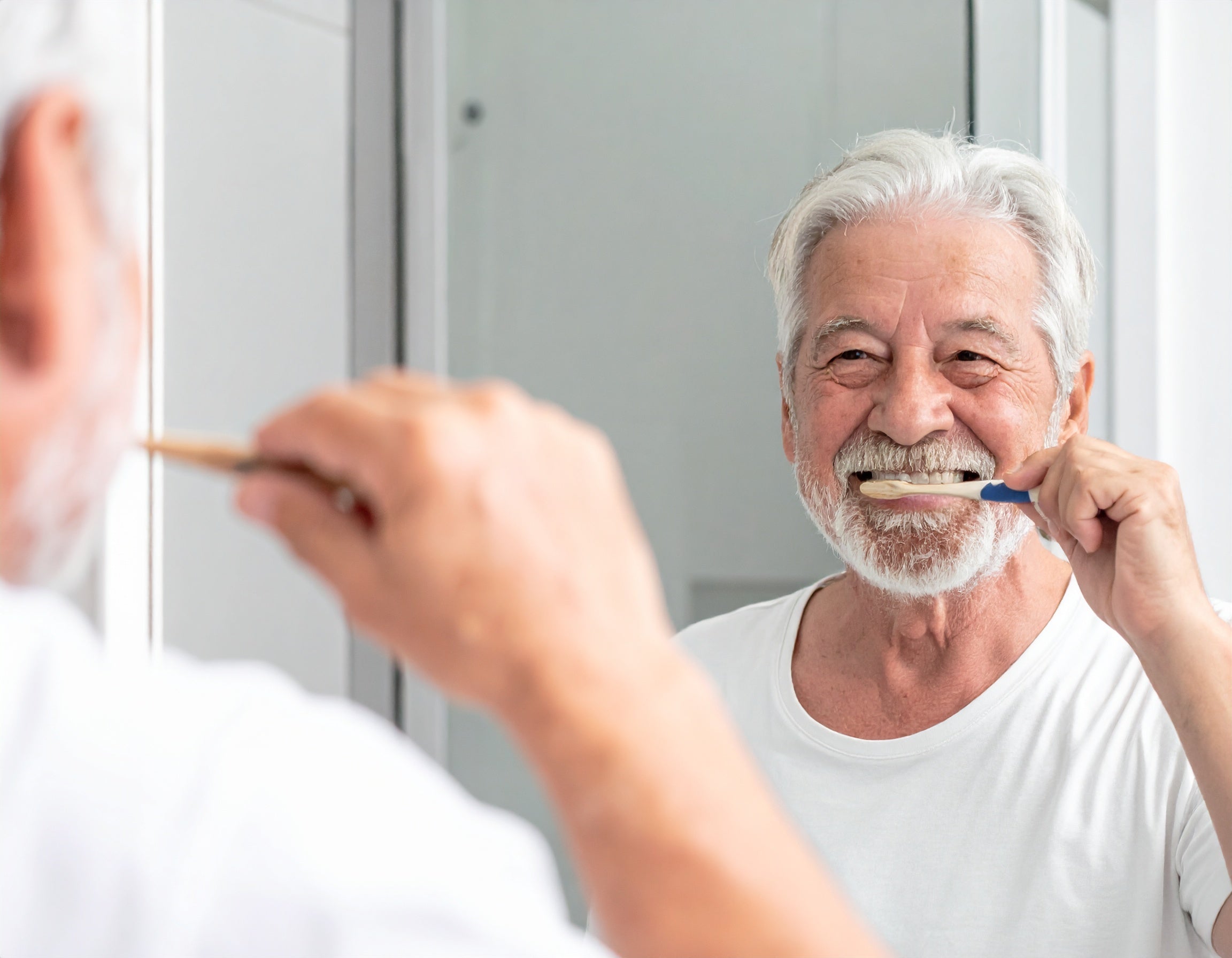 Na zdjęciu uśmiechnięty senior patrzący w lustro i myjący zęby szczoteczką.