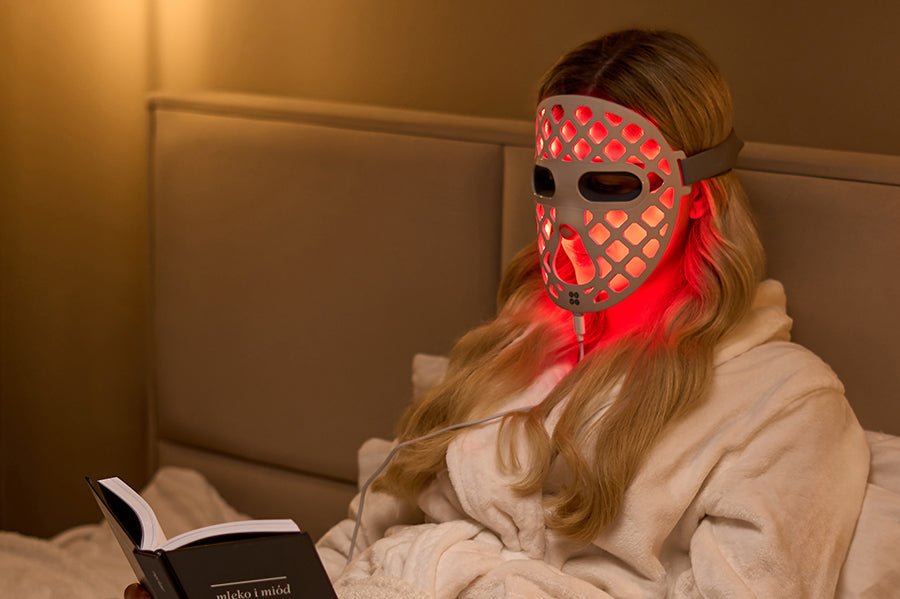 Na zdjęciu kobieta siedząca w łóżku i czytająca książkę. Na twarzy ma ZENT® DermaLight™ – Inteligentną maskę LED do fototerapii 4w1 w trybie światła czerwonego