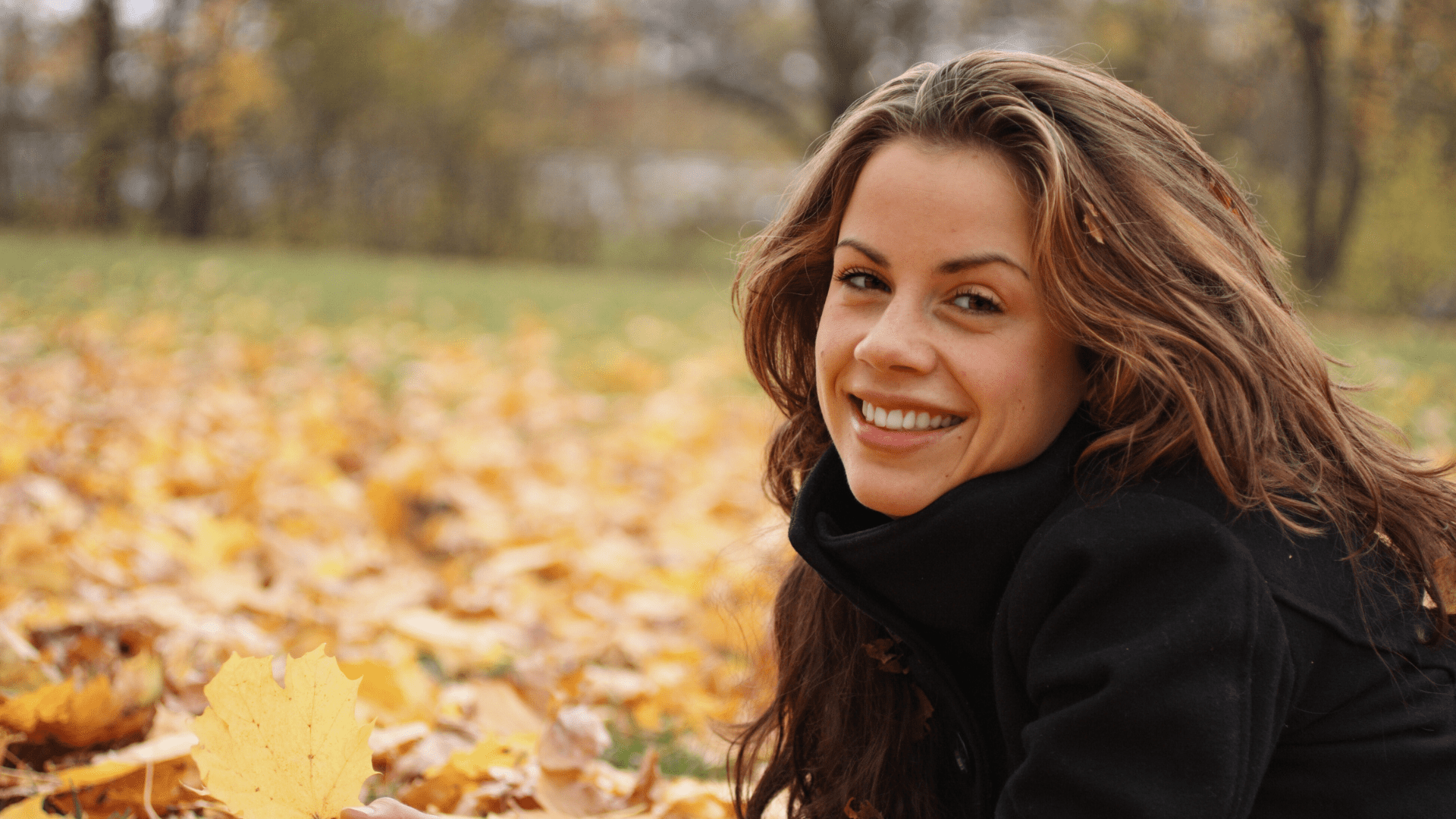 Uśmiechnięta kobieta wśród jesiennego pejzażu. Wpływ pór roku na zdrowie jamy ustnej, jak dbać o zęby jesienią?